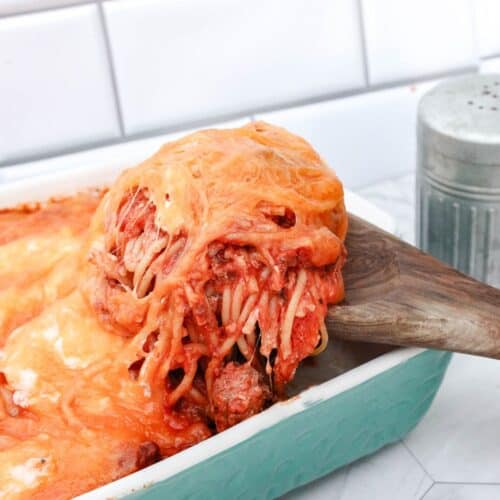 A wooden spatula lifts a serving of cheesy spaghetti with melted cheese from a turquoise casserole dish on a kitchen counter.