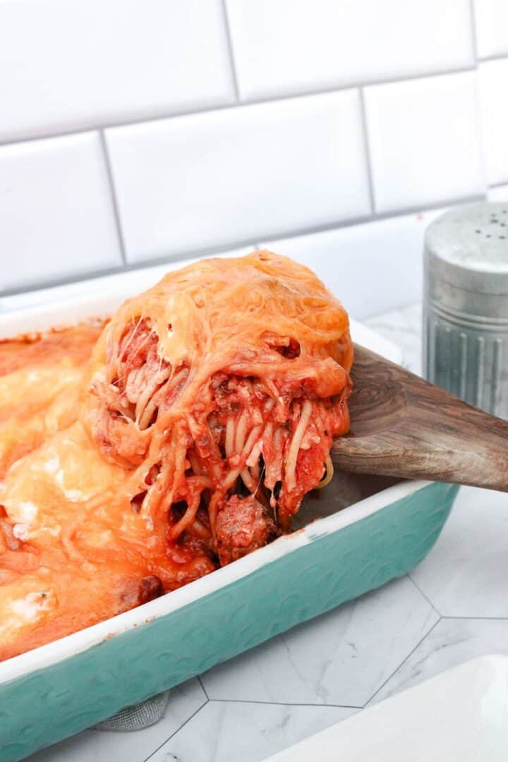 A wooden spatula lifts a serving of cheesy spaghetti with melted cheese from a turquoise casserole dish on a kitchen counter.