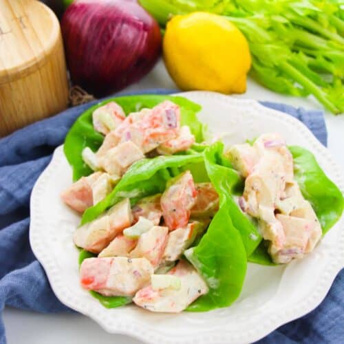 Plate of lettuce wraps filled with a creamy crab meat salad, surrounded by celery, a red onion, a lemon, and a pepper grinder on a white surface.