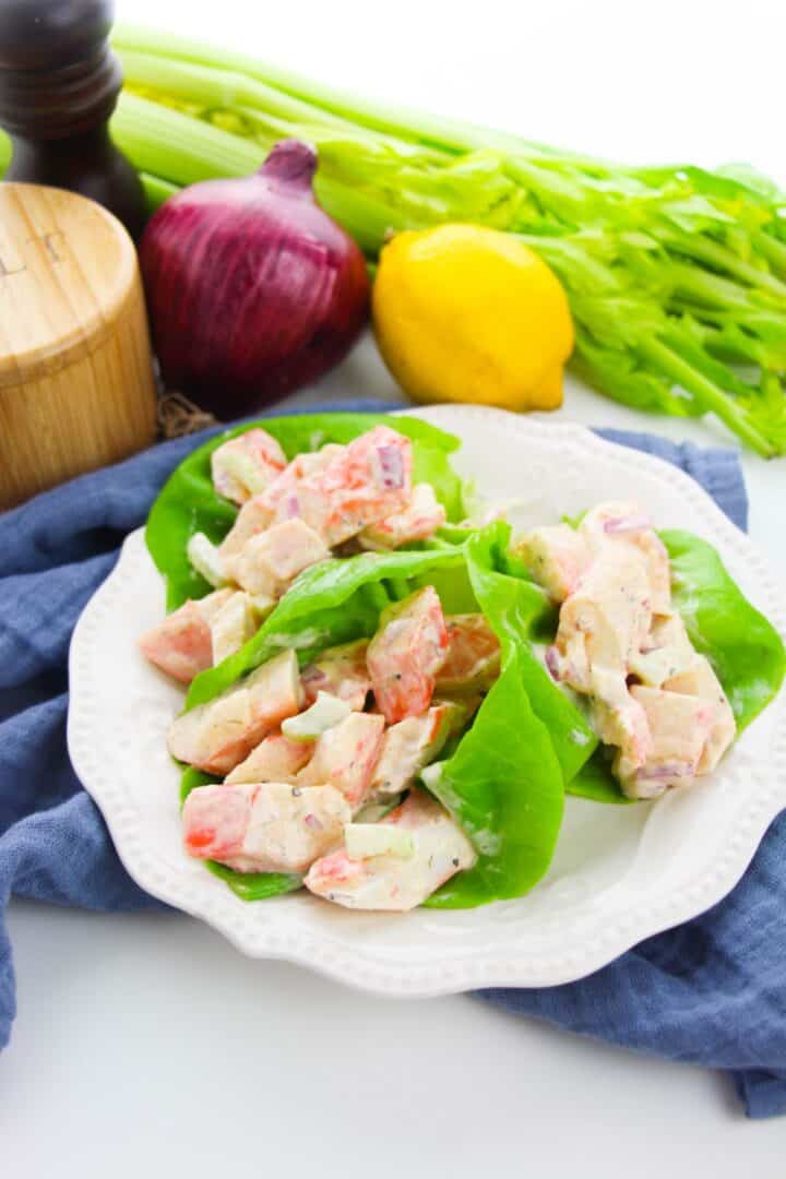Plate of lettuce wraps filled with a creamy crab meat salad, surrounded by celery, a red onion, a lemon, and a pepper grinder on a white surface.