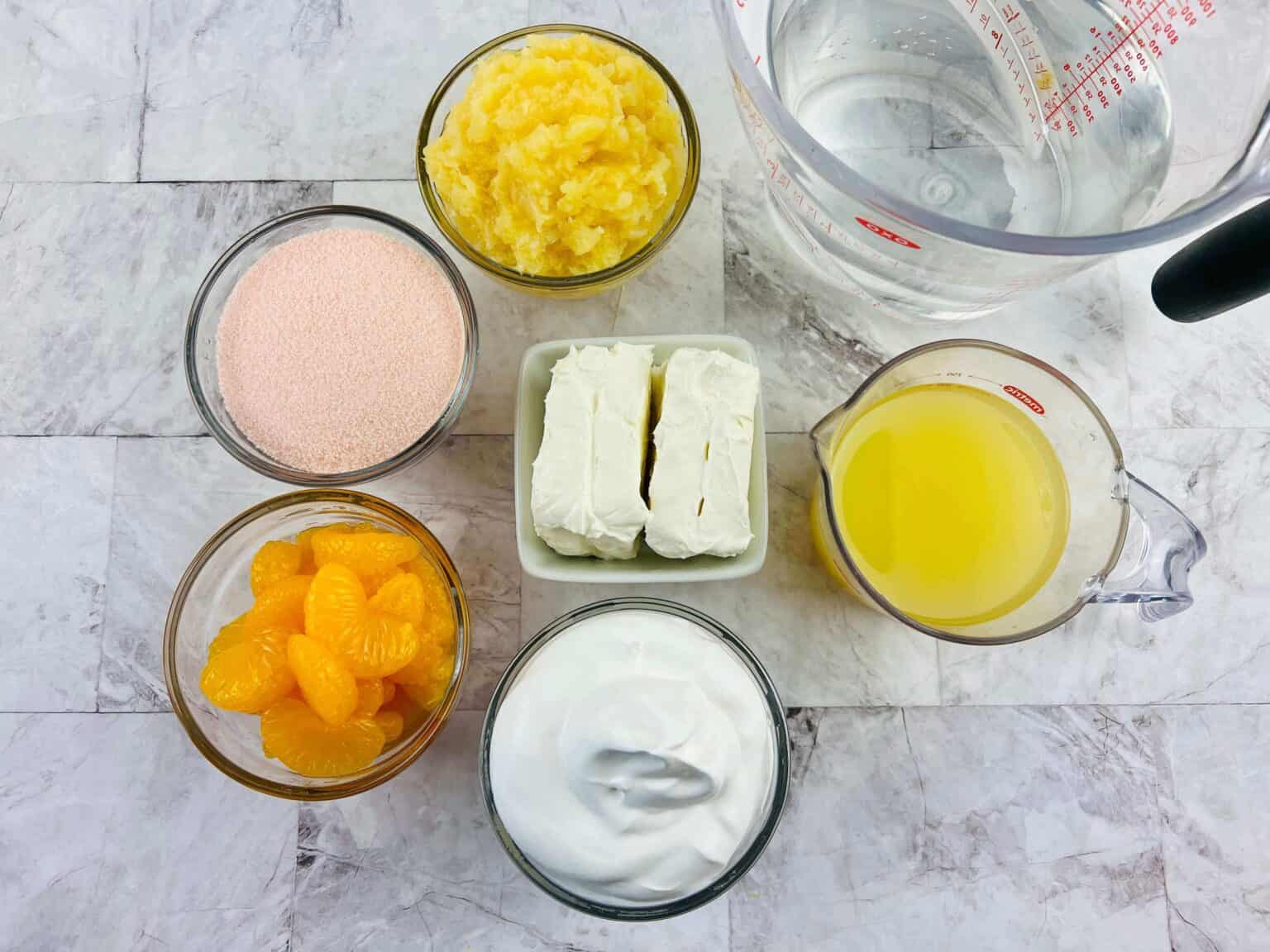 Ingredients for our Orange Jello Salad are artfully arranged on the table: crushed pineapple, sliced mandarins, orange powder, cream cheese, whipped topping, orange juice, and water.