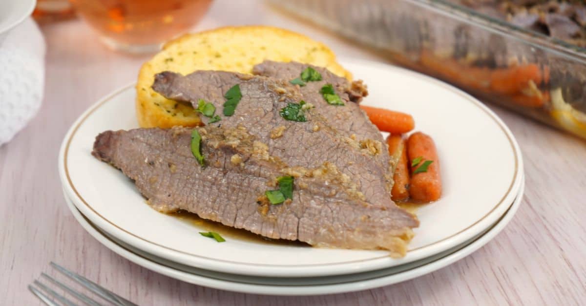 Two slices of oven brisket garnished with parsley, served alongside baby carrots and a slice of garlic bread on a white plate.