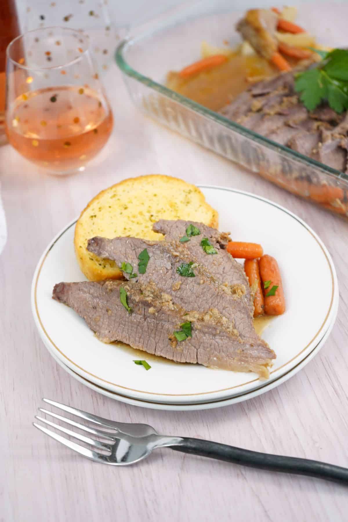 Sliced oven brisket with carrots and a piece of cornbread on a white plate, served alongside a glass of rosé and a dish of extra brisket in the background.