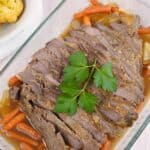 Sliced meat with carrots and onions in a glass baking dish, garnished with parsley. Garlic bread is visible on the side.