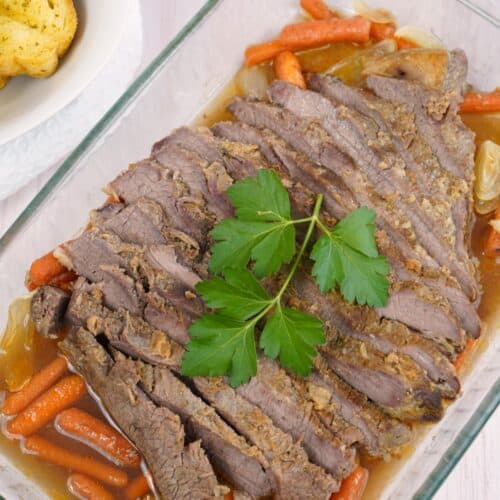 Sliced meat with carrots and onions in a glass baking dish, garnished with parsley. Garlic bread is visible on the side.