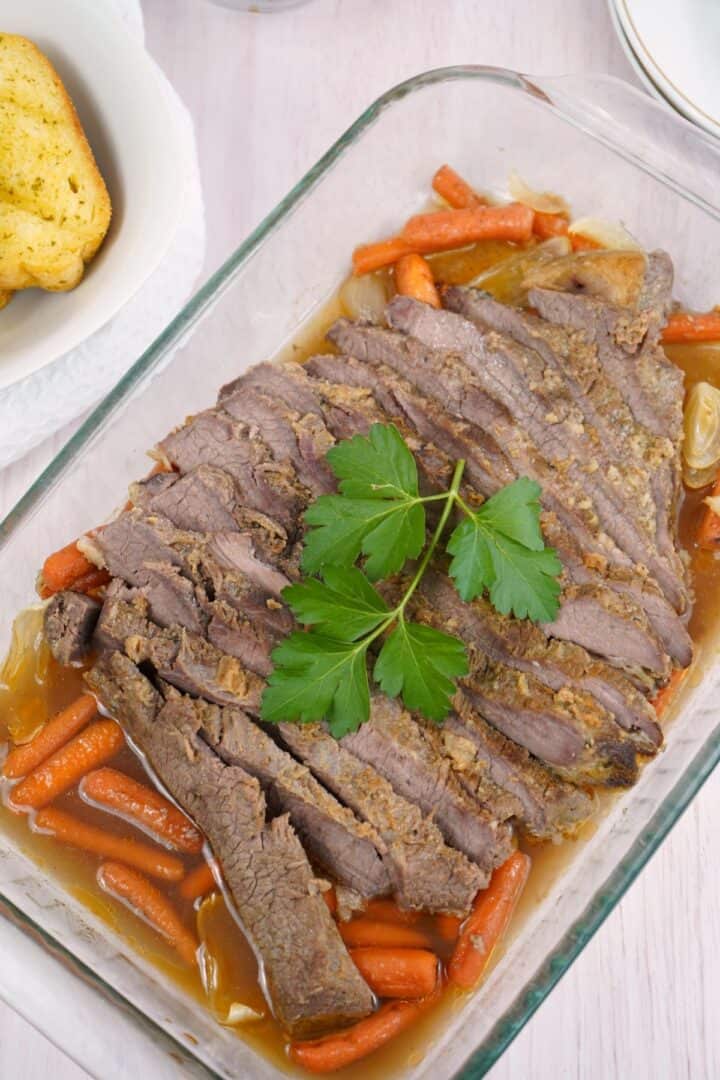 Sliced meat with carrots and onions in a glass baking dish, garnished with parsley. Garlic bread is visible on the side.