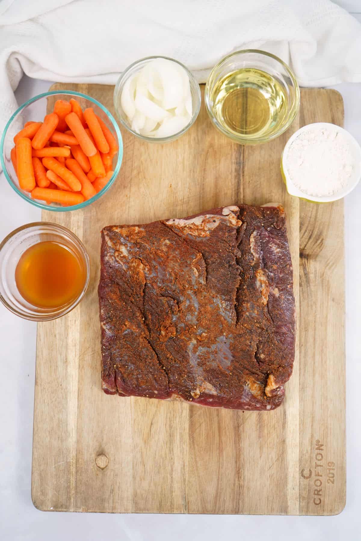 A wooden cutting board with a seasoned raw oven brisket, baby carrots, sliced onions, a bowl of flour, apple cider vinegar, and a cup of broth arranged on top.