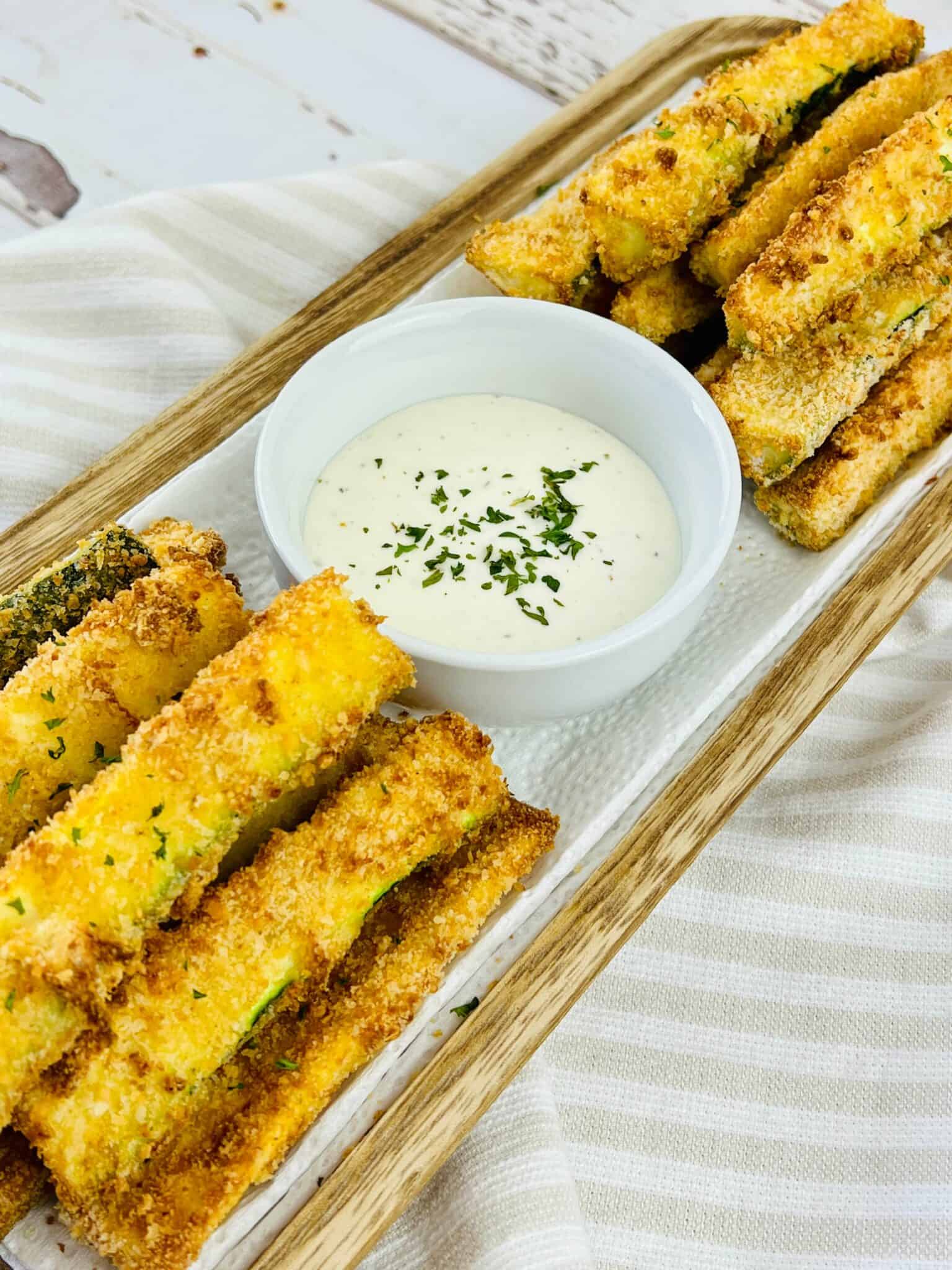 A wooden tray with the zucchini sticks and a bowl of dipping sauce in the middle.