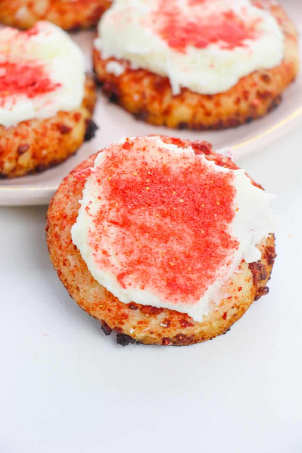 A Strawberry Crunch Cookie topped with white frosting and red sprinkles sits on a white surface, with more cookies on a plate in the background.