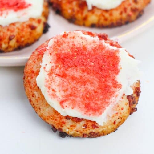 A Strawberry Crunch Cookie topped with white frosting and red sprinkles sits on a white surface, with more cookies on a plate in the background.
