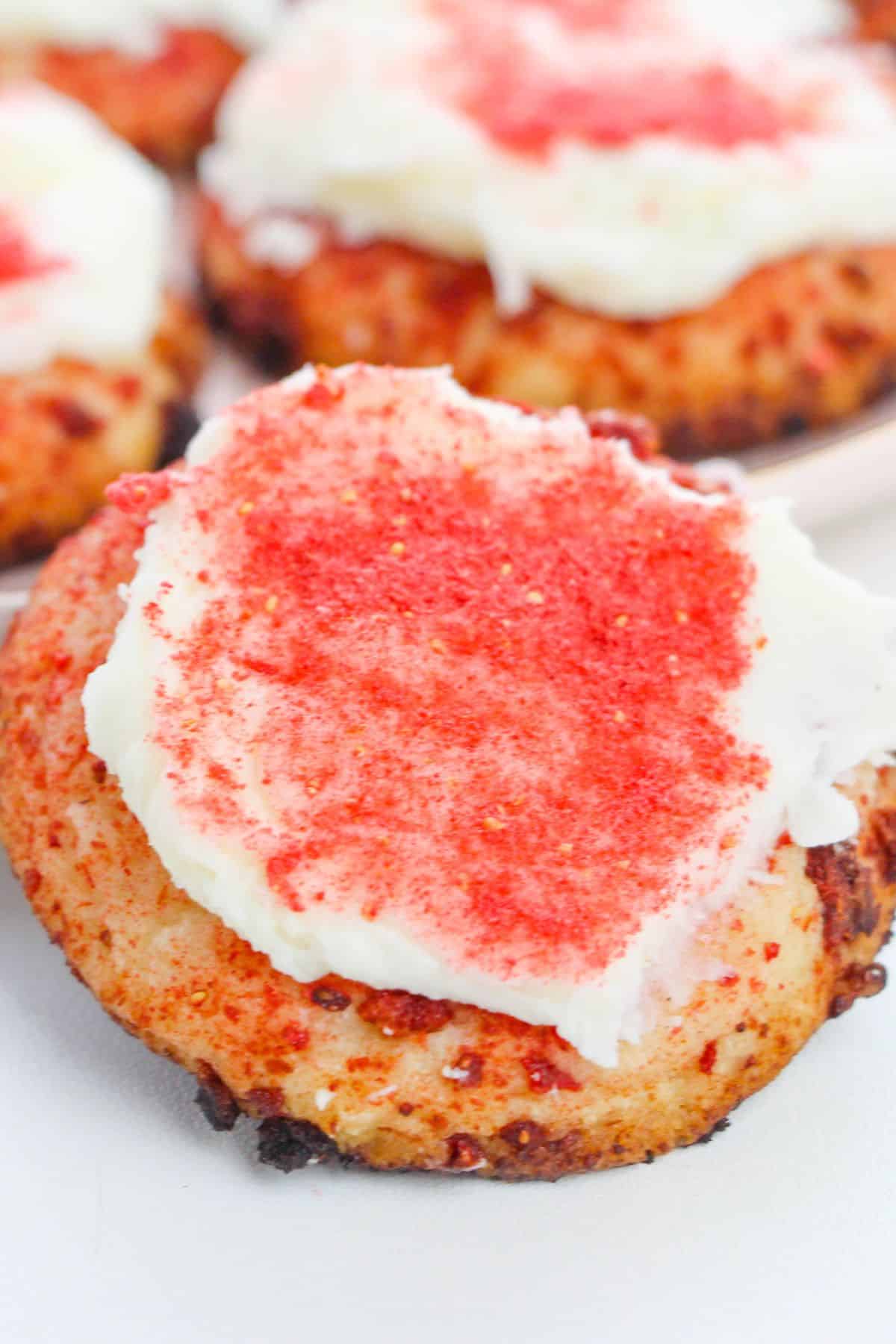 Close-up of a Strawberry Crunch Cookie topped with white frosting and red powder, with more frosted cookies in the background.