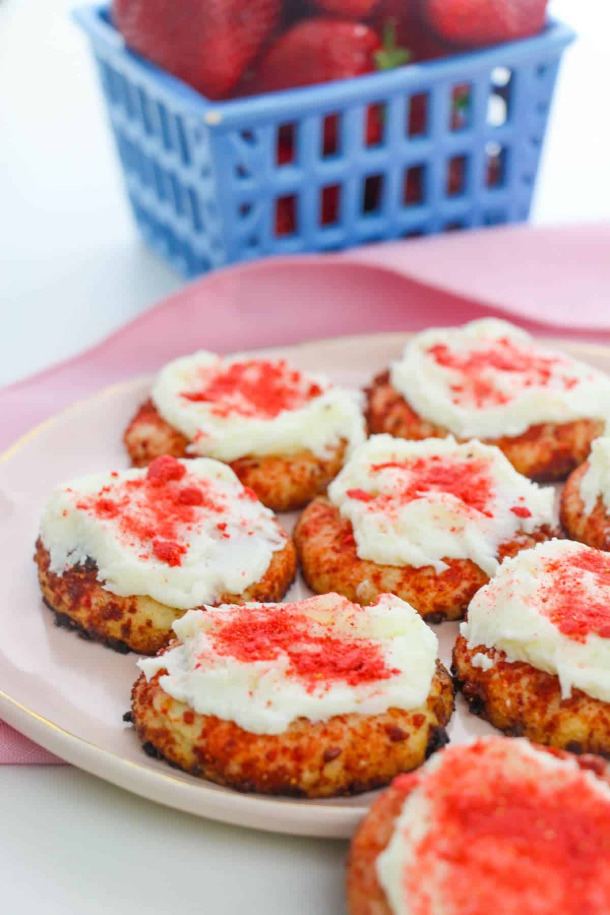A plate of the treats topped with white frosting and red sprinkles, with a blue basket of strawberries in the background.