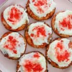 A plate of eight round Strawberry Crunch Cookies topped with white frosting and red sprinkles, arranged neatly on a light pink plate.