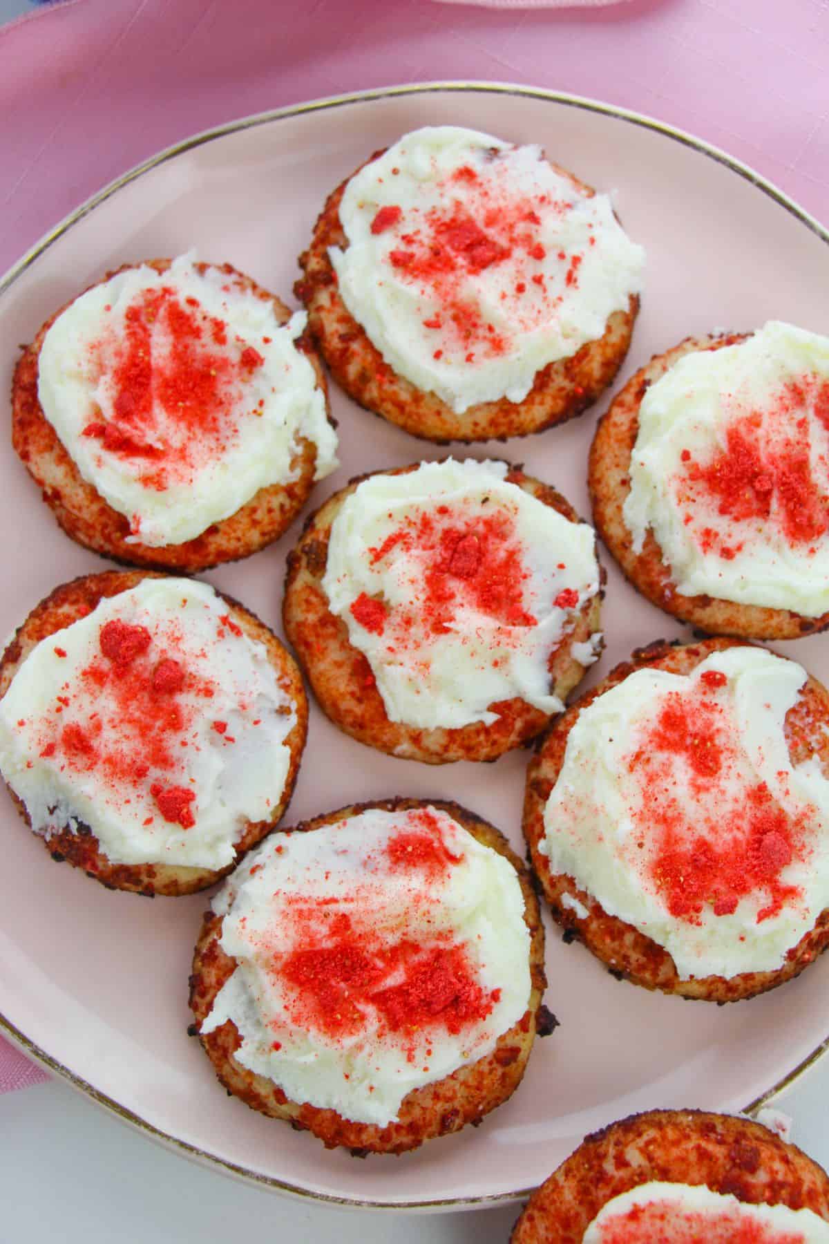 A plate of eight round Strawberry Crunch Cookies topped with white frosting and red sprinkles, arranged neatly on a light pink plate.
