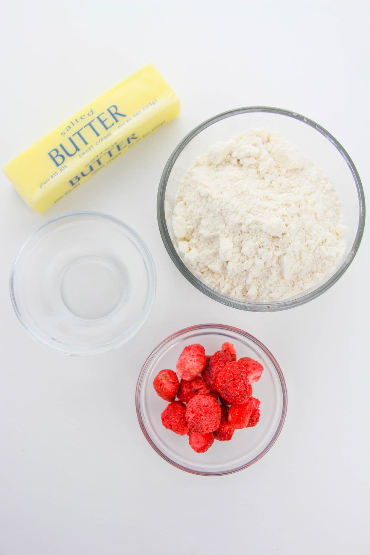 A stick of salted butter, a bowl of flour, a bowl of water, and a bowl of frozen strawberries are arranged on a white surface—perfect for whipping up homemade Strawberry Crunch Cookies.