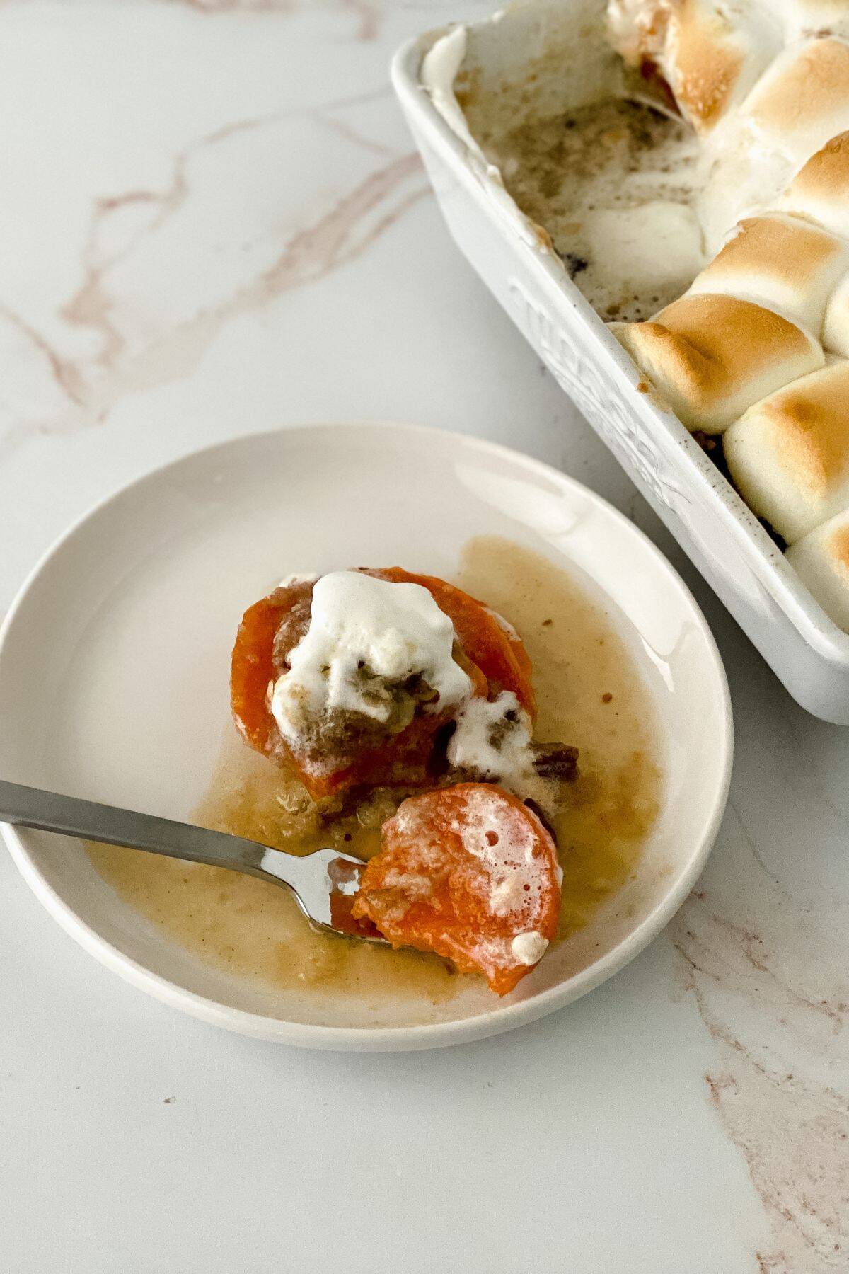 A serving of Sweet Potato Crunch casserole topped with toasted marshmallows on a white plate, with a fork and the casserole dish in the background.