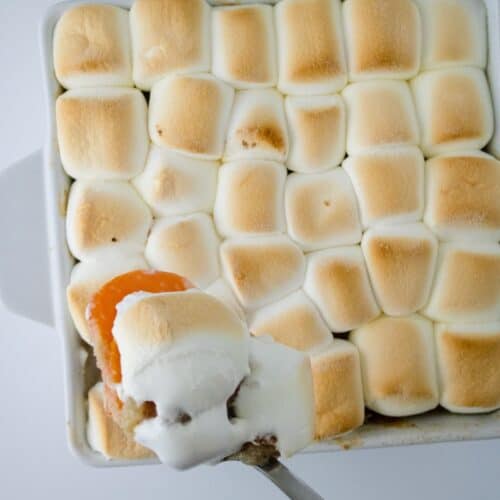 A hand holds a serving spoon with a portion of Sweet Potato Crunch casserole, topped with toasted marshmallows, taken from a baking dish.