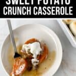 A serving of Sweet Potato Crunch casserole topped with cream sits on a white plate beside a fork. In the upper right corner, part of the Sweet Potato Crunch casserole dish is also visible.