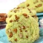 A close-up of a light green Pistachio Pudding Cookie topped with chopped pecans, placed on a blue plate with more cookies and pecans in the background.