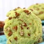 A light green Pistachio Pudding Cookie with chopped pecans on top, placed on a blue plate with other similar cookies in the background.