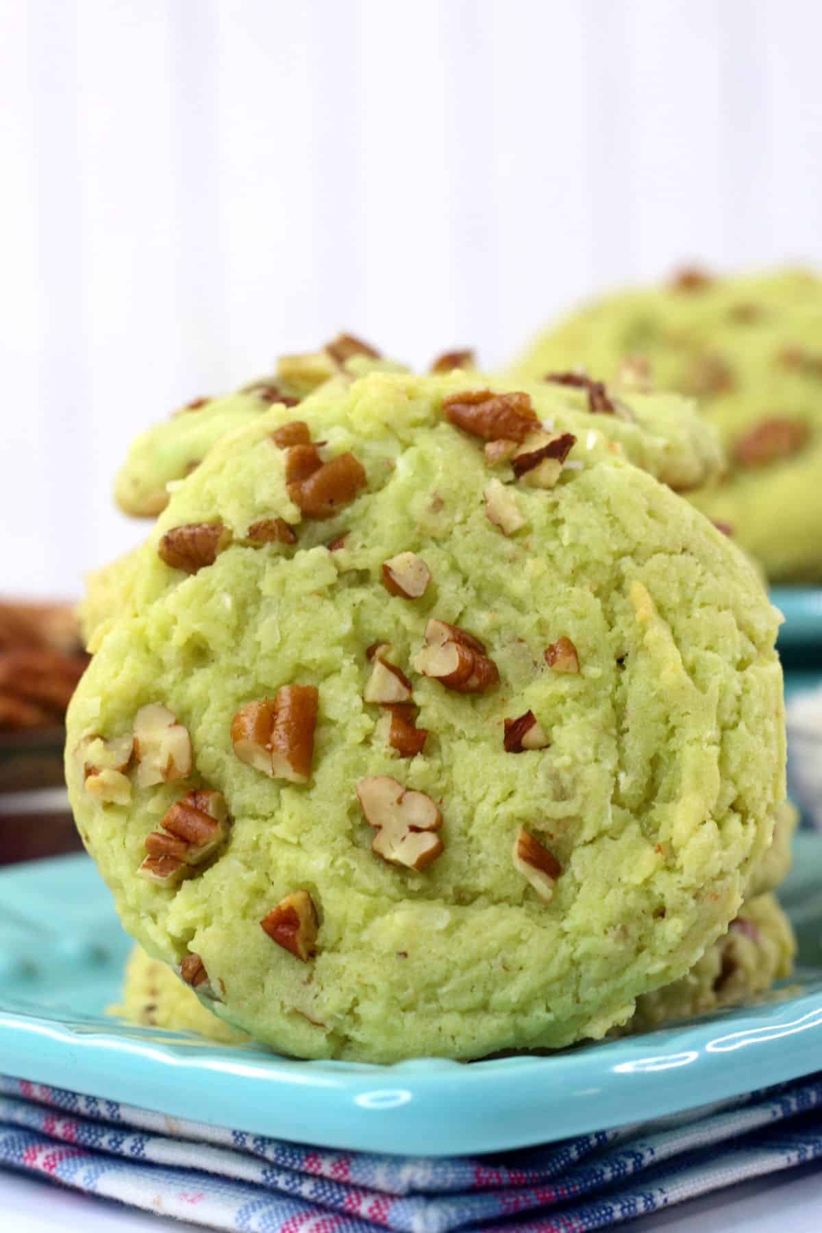 A light green Pistachio Pudding Cookie with chopped pecans on top, placed on a blue plate with other similar cookies in the background.