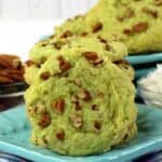 A stack of green Pistachio Pudding Cookies with chopped pecans sits on a blue plate, alongside bowls of pecans and shredded coconut.