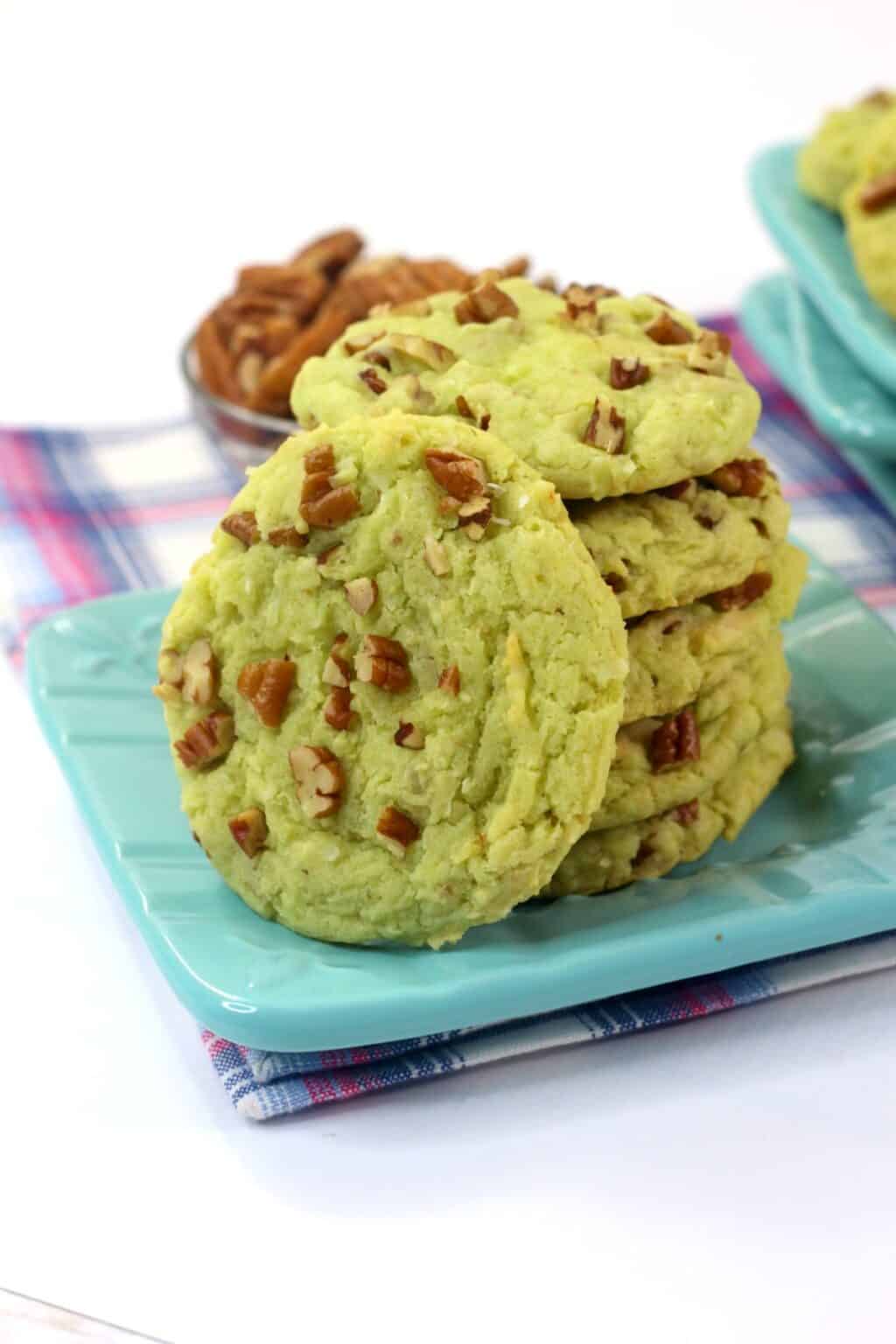 A stack of green Pistachio Pudding Cookies with chopped nuts sits on a blue plate, while more cookies and nuts are scattered in the background on a checkered cloth.