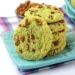 A stack of green Pistachio Pudding Cookies with chopped nuts sits on a blue plate, while more cookies and nuts are scattered in the background on a checkered cloth.
