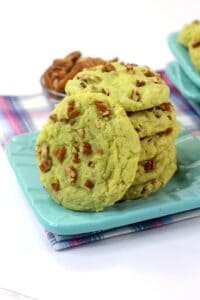 A stack of green Pistachio Pudding Cookies with chopped nuts sits on a blue plate, while more cookies and nuts are scattered in the background on a checkered cloth.