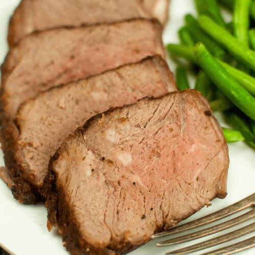 Sliced medium-rare Air Fryer Beef Roast arranged on a white plate with a side of green beans and a silver fork.