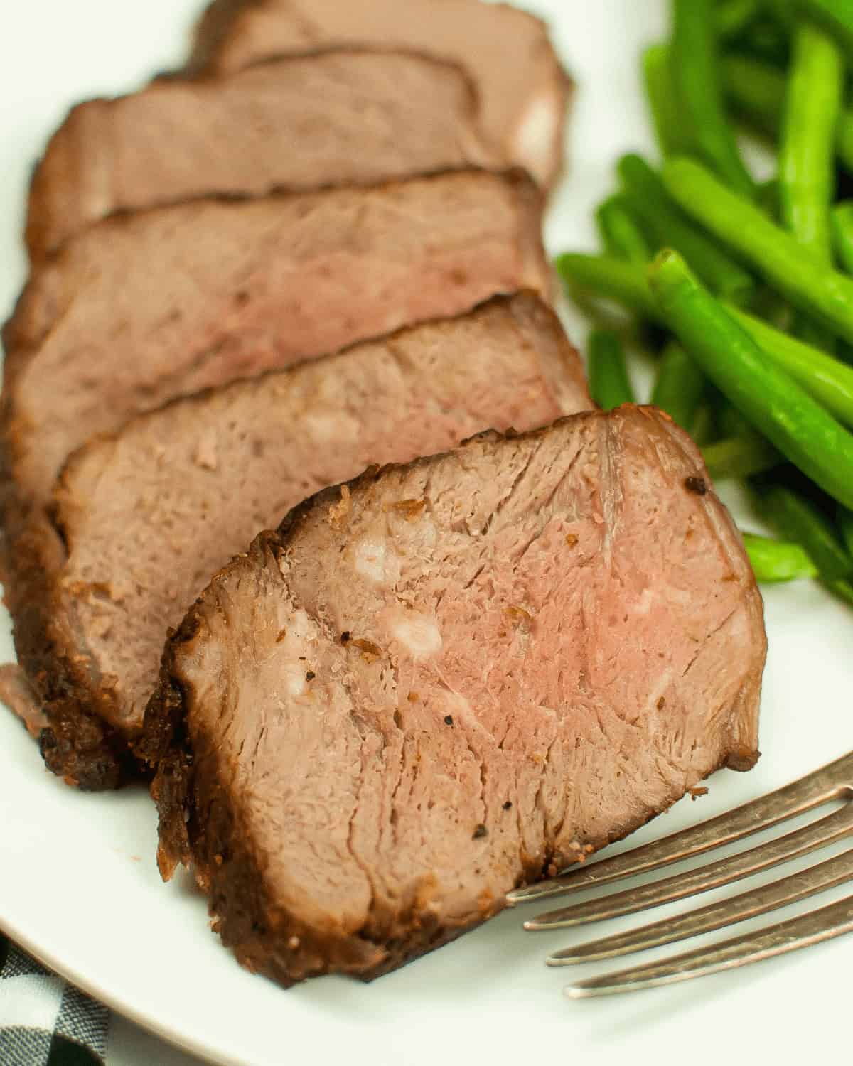 Sliced medium-rare Air Fryer Beef Roast arranged on a white plate with a side of green beans and a silver fork.