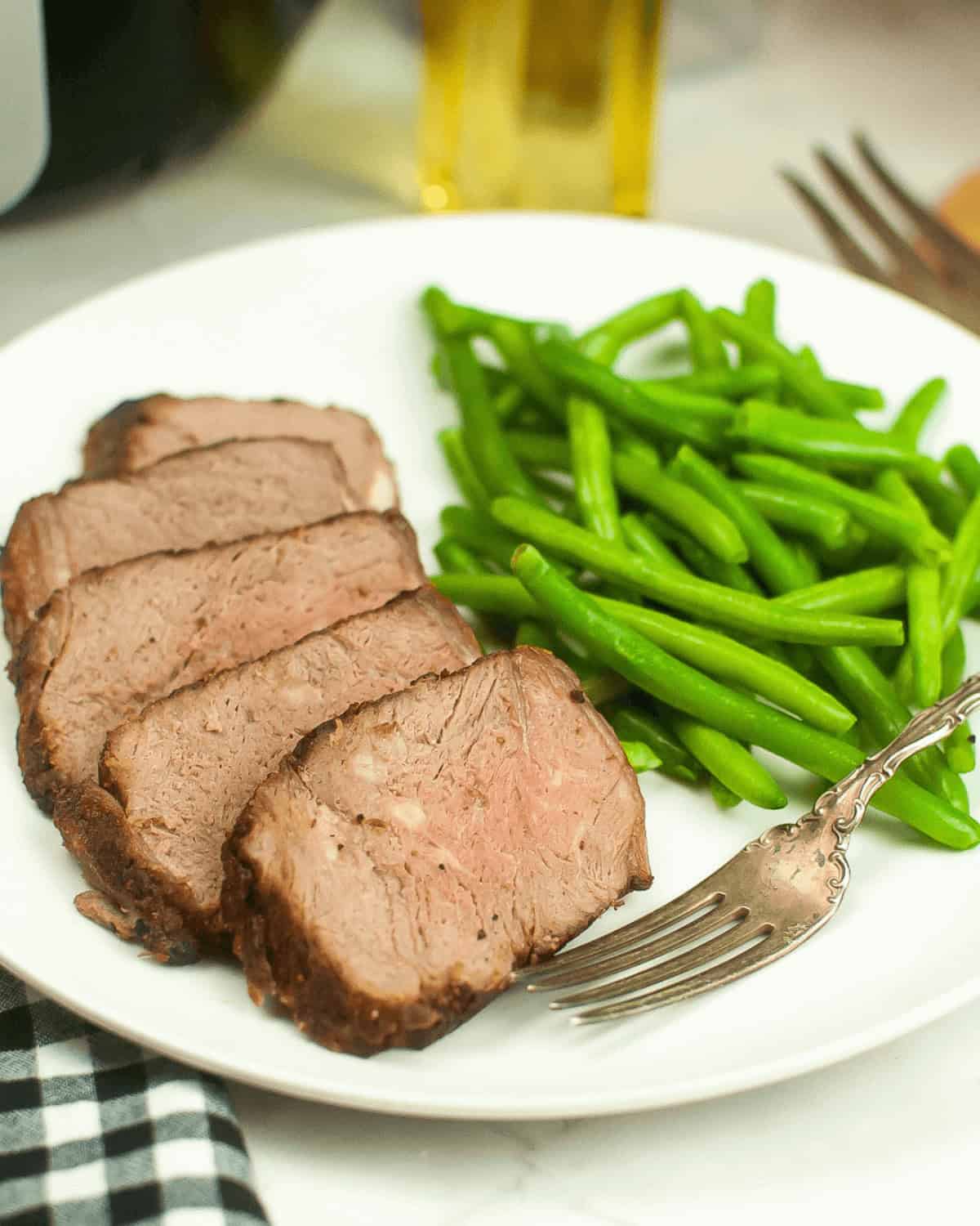 Sliced Air Fryer Beef Roast and steamed green beans arranged on a white plate with a fork, set on a table with a checkered napkin.