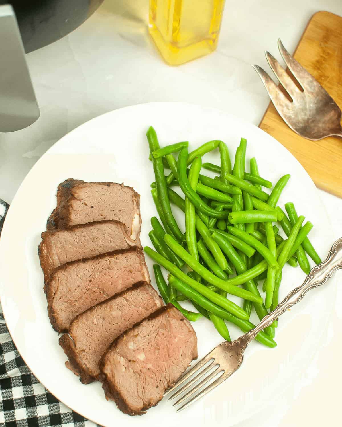 A white plate with four slices of tender Air Fryer Beef Roast and a serving of green beans, with a fork on the plate and utensils on a wooden board nearby.