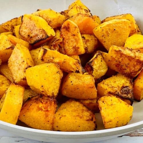 A white bowl filled with roasted, seasoned Air Fryer Butternut Squash cubes sits on a white surface with a spoon nearby.