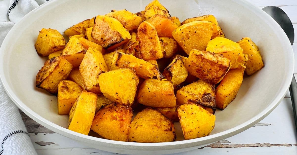A white bowl filled with roasted, seasoned Air Fryer Butternut Squash cubes sits on a white surface with a spoon nearby.