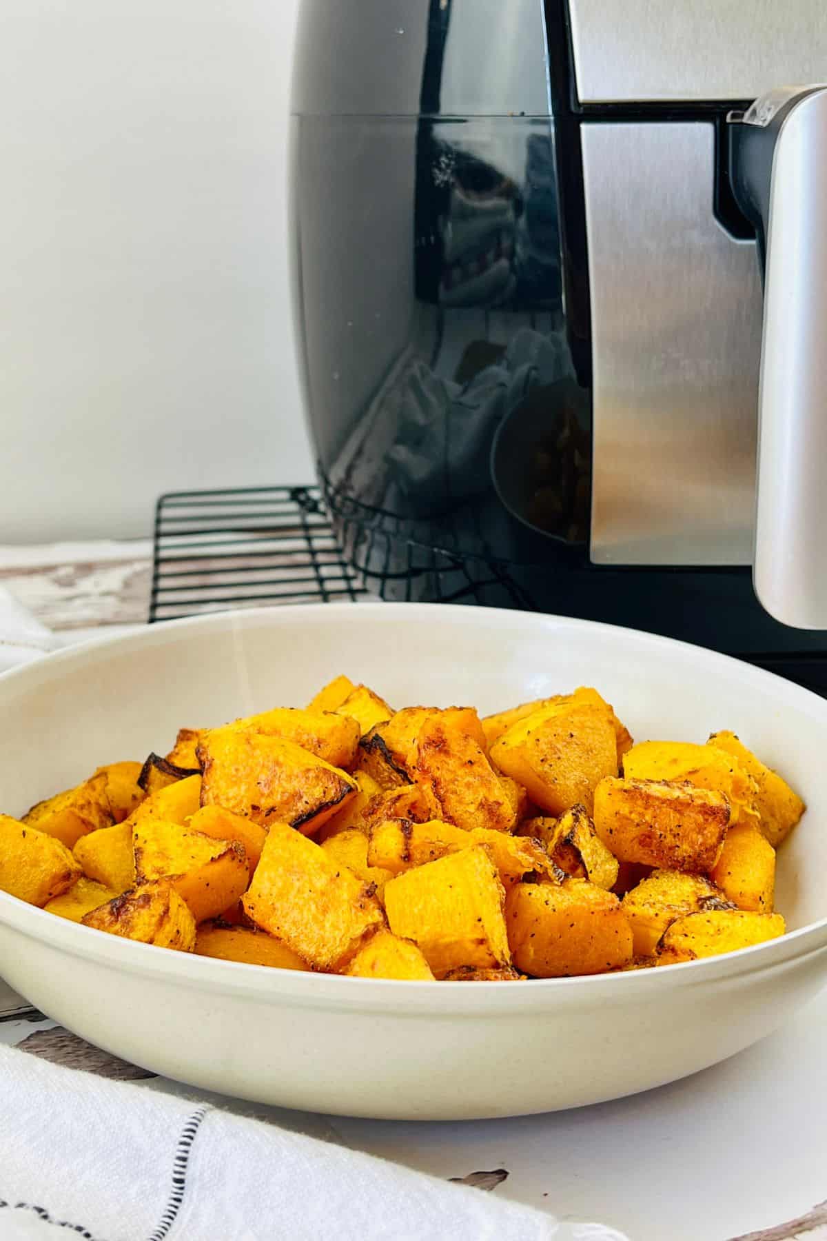 A white bowl filled with perfectly roasted Air Fryer Butternut Squash cubes sits in front of a sleek black air fryer on a countertop.