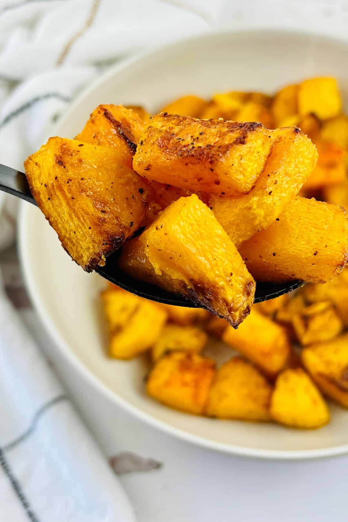 A spoon holds golden brown, perfectly seasoned Air Fryer Butternut Squash cubes, with more roasted squash in a bowl in the background.