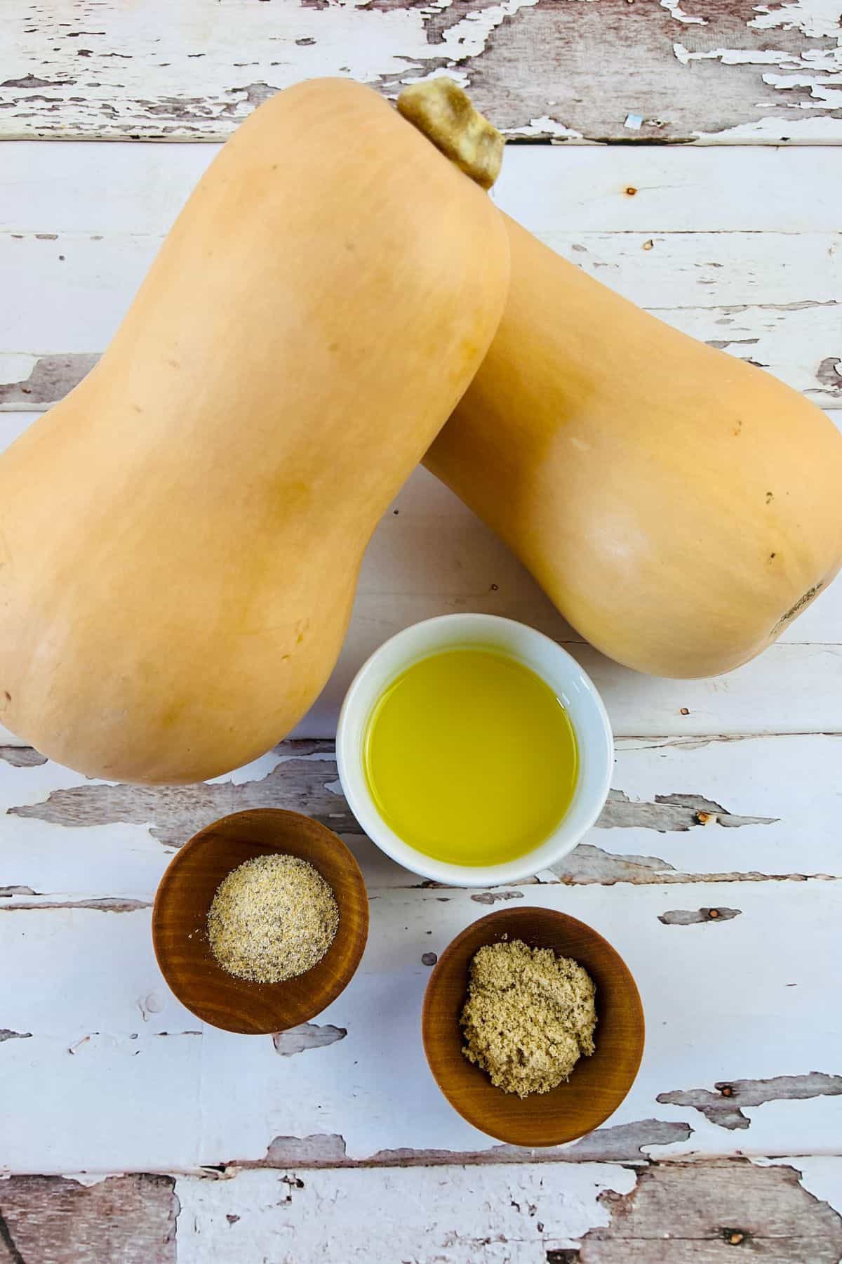 Two whole butternut squash, a small bowl of olive oil, a bowl of ground pepper, and a bowl of brown sugar are arranged on a rustic white wooden surface—perfect ingredients to create delicious Air Fryer Butternut Squash.