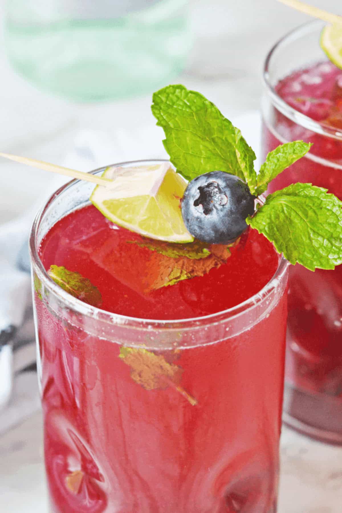 A glass of red beverage, reminiscent of a Blueberry Mojito, is garnished with a skewer holding a blueberry, mint leaves, and a lime wedge. Another similar drink graces the background.