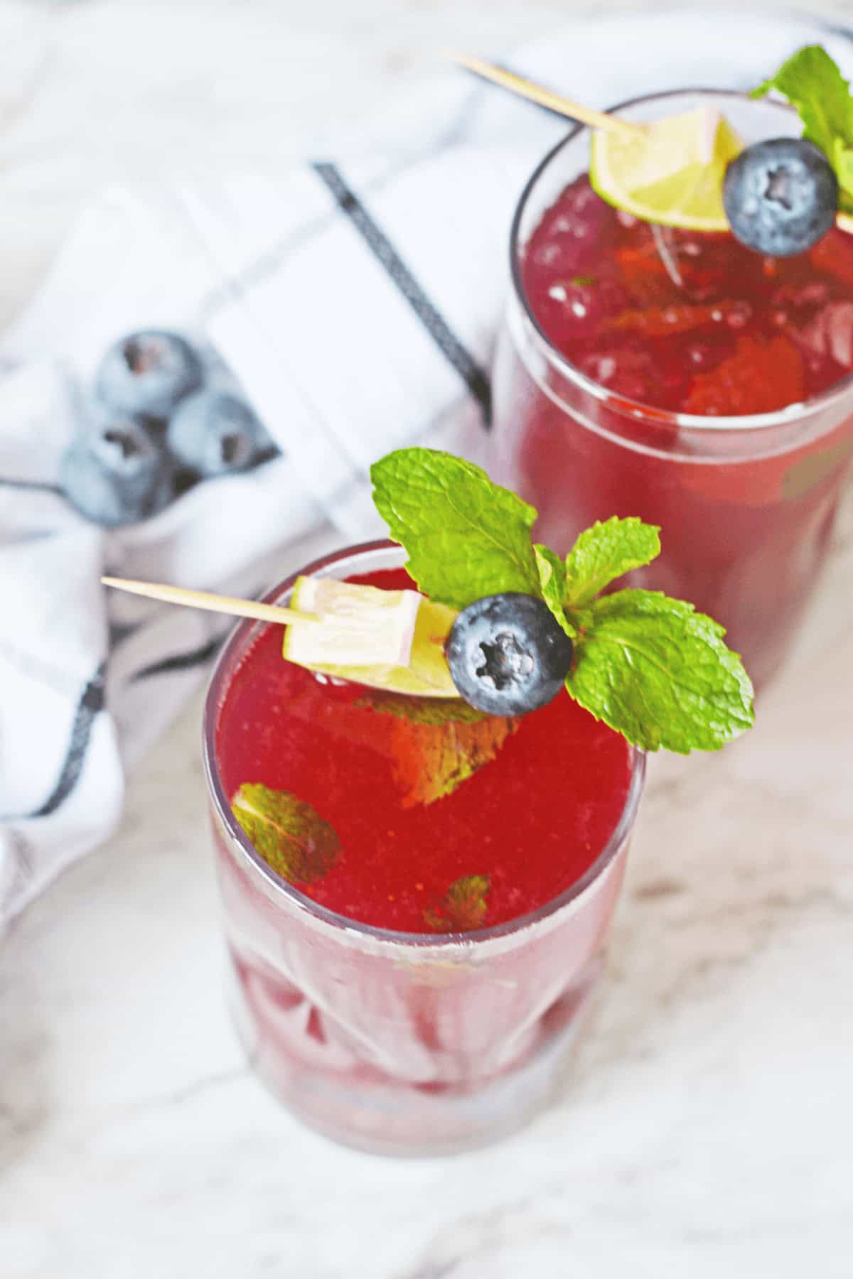 Two glasses of Blueberry Mojito garnished with mint leaves, blueberries, and lemon wedges on a white surface. A few blueberries and a cloth napkin are in the background.