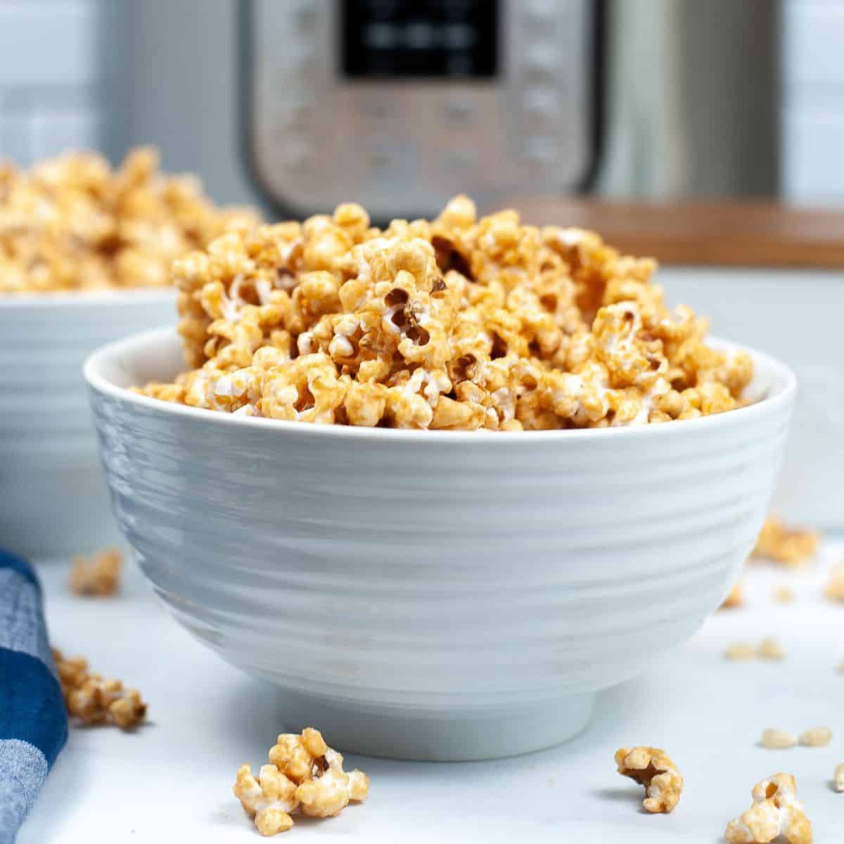 A white bowl filled with caramel-coated Instant Pot Popcorn sits on a white surface with a few pieces scattered around.