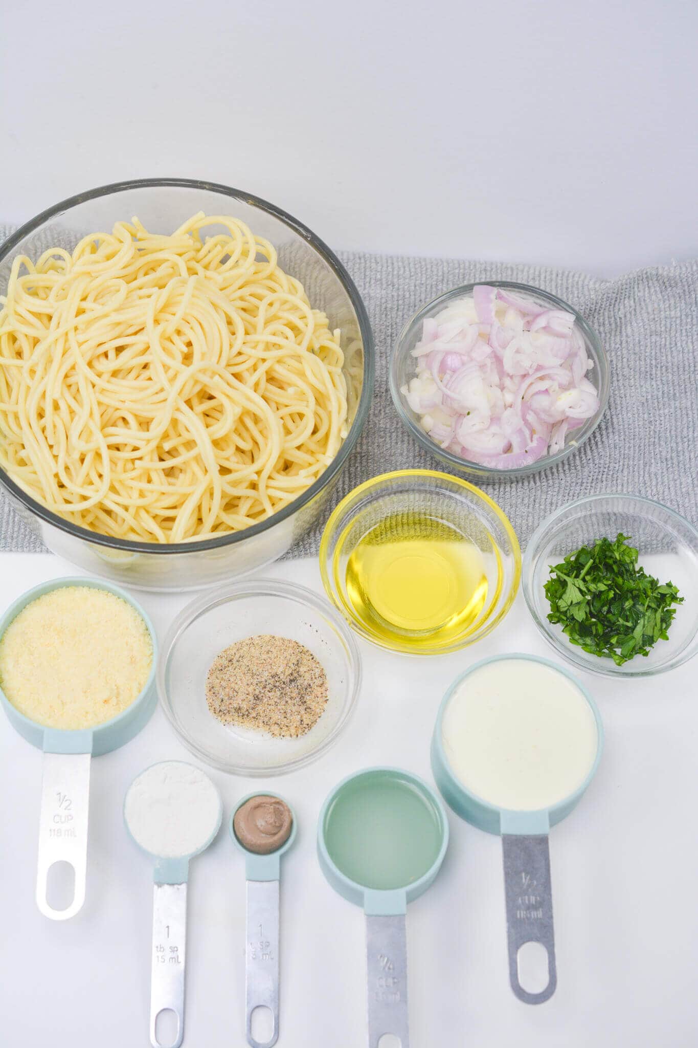 A bowl of cooked spaghetti, caramelized onion pasta, olive oil, chopped parsley, grated cheese, spices, flour, mustard, water, and cream in measuring cups rest on a gray cloth.