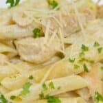 Penne pasta with chunks of chicken gorgonzola in a creamy sauce, topped with shredded cheese and chopped parsley, served on a white plate.