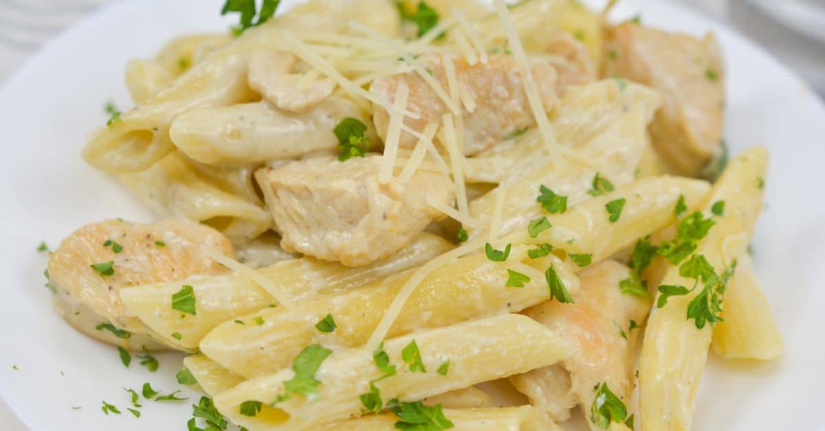 Penne pasta with chunks of chicken gorgonzola in a creamy sauce, topped with shredded cheese and chopped parsley, served on a white plate.