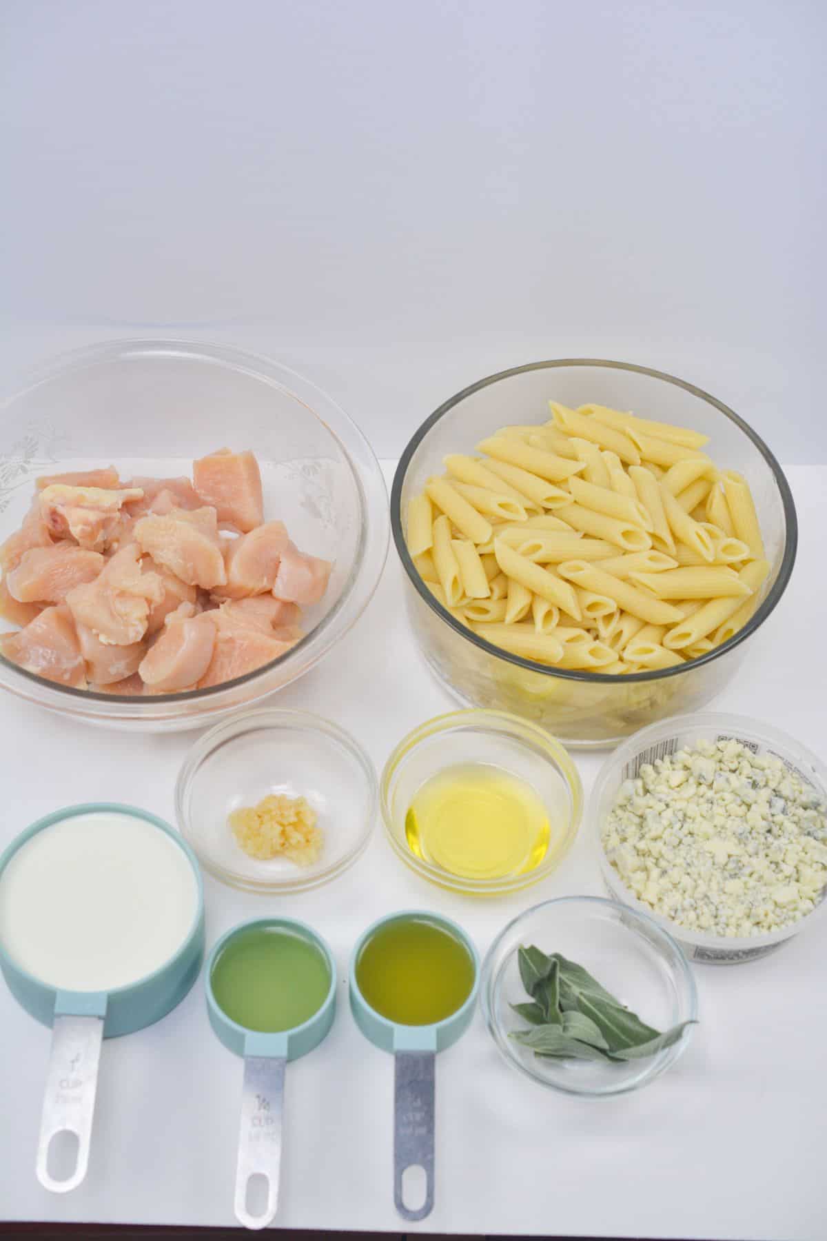 Bowls of uncooked penne pasta, cubed raw chicken, cream, chopped garlic, oil, crumbled Gorgonzola cheese, fresh sage, and seasonings arranged on a white surface—everything you need to create a savory Chicken Gorgonzola pasta dish.
