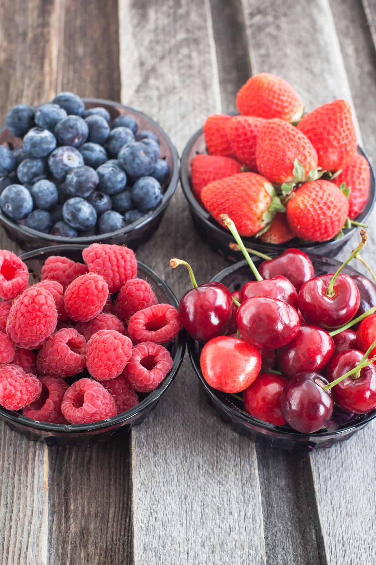 Four glass bowls filled with blueberries, strawberries, raspberries, and cherries are arranged on a wooden surface, perfect for creating a vibrant Strawberry Blueberry Salad.