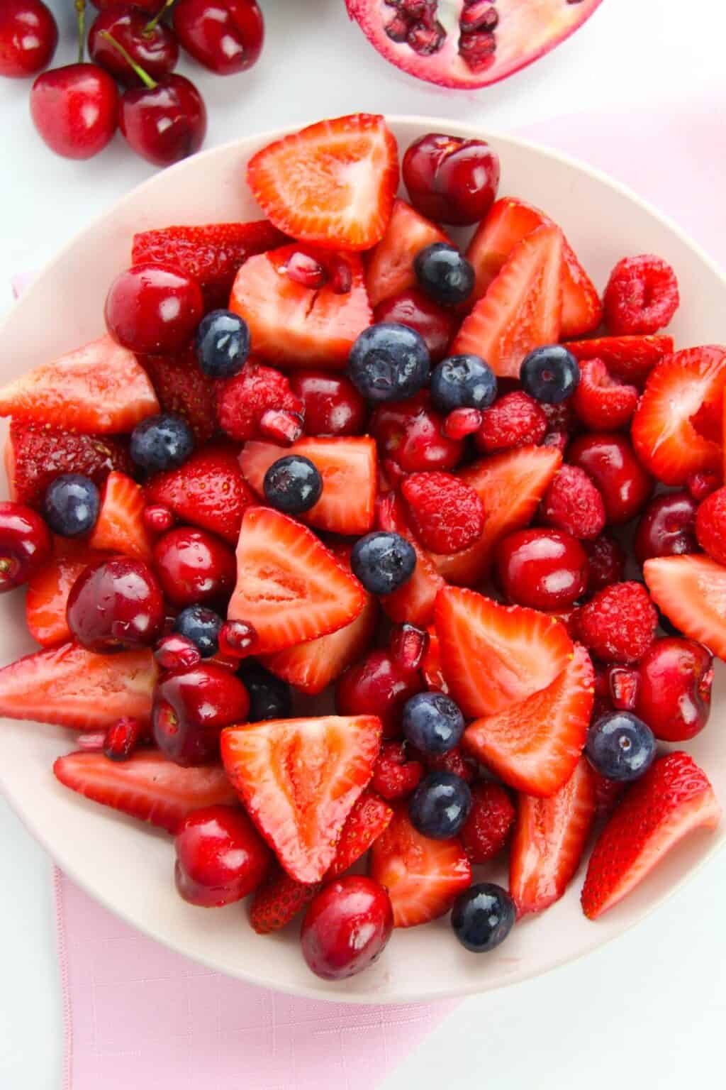 A vibrant Strawberry Blueberry Salad featuring fresh strawberries, blueberries, cherries, raspberries, and pomegranate seeds rests on a light pink surface.