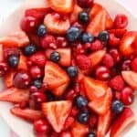 A vibrant Strawberry Blueberry Salad featuring fresh strawberries, blueberries, cherries, raspberries, and pomegranate seeds rests on a light pink surface.
