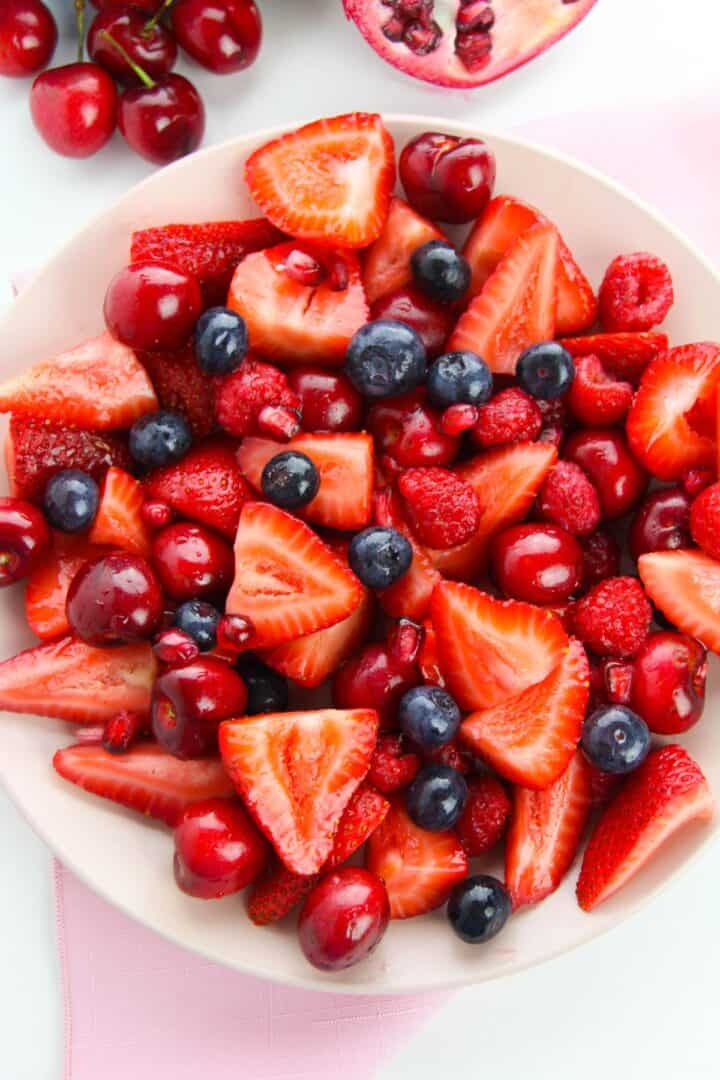 A vibrant Strawberry Blueberry Salad featuring fresh strawberries, blueberries, cherries, raspberries, and pomegranate seeds rests on a light pink surface.
