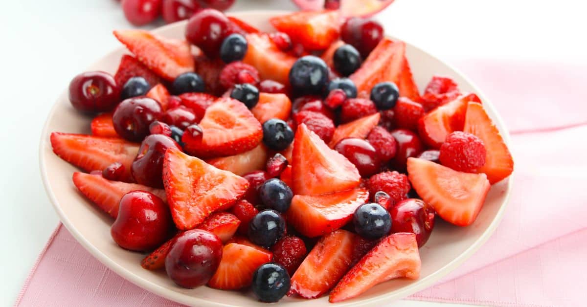 A plate filled with sliced strawberries, whole blueberries, cherries, and raspberries on a pink napkin makes a colorful Strawberry Blueberry Salad perfect for any occasion.